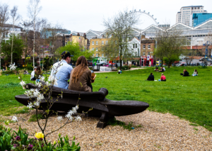 Two people are seated on a bench, in a park. The image looks at their back. On the background, some houses and a Ferris wheel