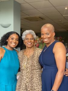 Three women of varying ages (three different generations) stand, smiling at the camera