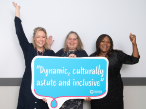 Three happy women behind a sign with the words "Dynamic, culturally astute and inclusive"
