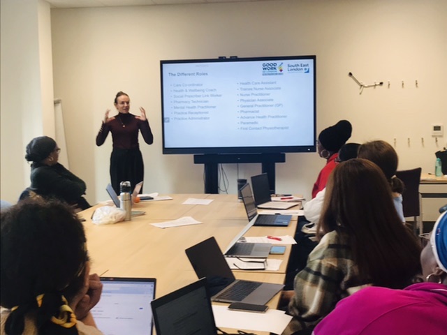 Someone delivering the SWAP scheme tutorial at the Tessa Jowell Health Centre: a person is talking near to a big TV screen, delivering the training to a series of people seated around a table