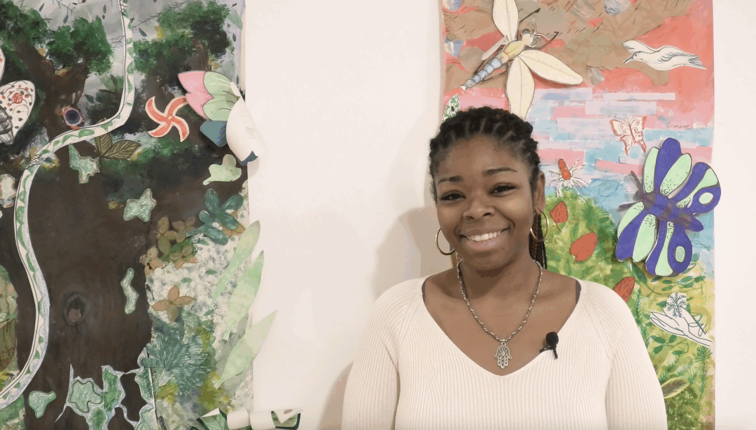 A person wearing a light-colored top and a necklace with a pendant is standing in front of two colorful, nature-themed artworks. The artworks feature butterflies, dragonflies, leaves, and abstract shapes in vibrant greens, reds, and blues.