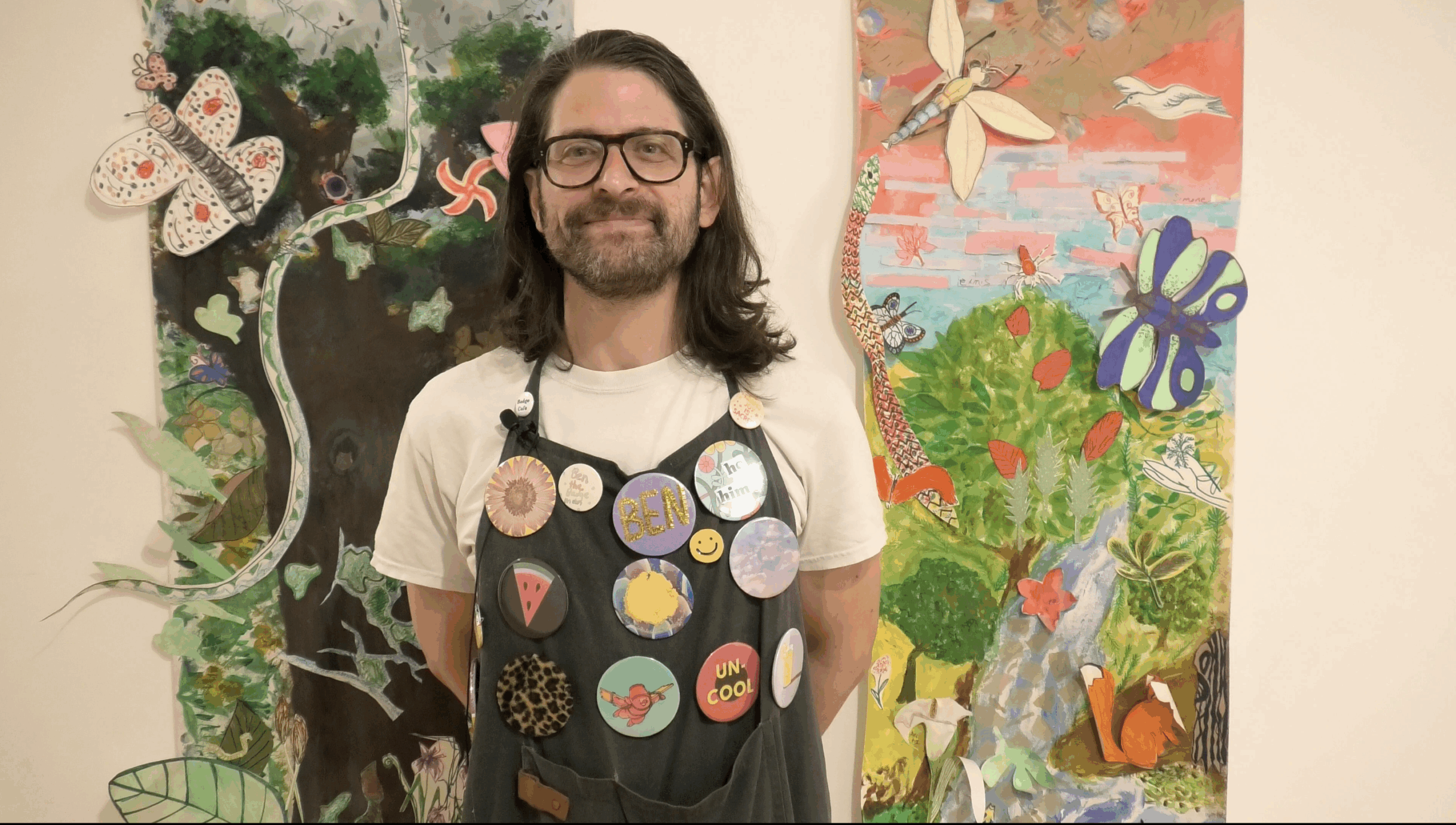 A person wearing a white shirt and a dark apron decorated with multiple colorful badges is standing in front of the same two nature-themed artworks. The badges include various designs such as smiley faces, text, and patterns.