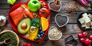 Image of a variety of healthy foods on a red, heart-shaped plate. These foods include salmon, carrots, apples, tomatoes, blueberries, olive oil, beans, avocados and salmon. Surrounding this plate, smaller bowls with seeds in them.
