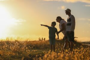Eye-level view of a family silhouetted against a vibrant sunset. The family consists of a man, a woman, and a young boy. The man is wearing a bucket hat and what appears to be a striped, short-sleeved shirt and shorts. The woman is wearing a sleeveless top and is holding the boy's hand. The boy is pointing towards the horizon. They are standing in a field of tall, golden-colored grass, which extends to the horizon. The sunset is a warm, rich orange and yellow, with a slightly hazy sky. The overall mood is serene and peaceful, suggestive of a family enjoying a moment together in nature. The silhouettes are sharply defined against the bright background, emphasizing their connection and the beautiful setting