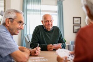 Four seniors are seated around a table, playing cards and engaged in conversation. The central figure, a man with a dark green sweater, holds a hand of cards, smiling at his companions. The atmosphere is relaxed and friendly.
