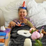 Close-up view of a woman lying in a hospital bed, wearing a Union Jack party hat and a striped shirt. She's smiling broadly and holding a plate with what appears to be a piece of cake, decorated with a Union Jack design. She's using a utensil to eat the cake. The bed is made with white linens and a gray blanket is draped over her. A bedside table is partially visible, holding tissues and other items. Pink peonies in a vase are in the foreground, slightly out of focus