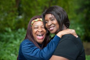 Two women stand outside in a green, leafy setting, smiling and embracing. The woman on the left is laughing with her mouth open wide, wearing a colourful headband and a long-sleeved blue top. Her arms are around the other woman, who has shoulder-length dark hair and wears a black top. They look relaxed, happy and close, enjoying a light moment together.