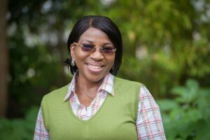 A medium close-up shot portrays a Black woman with a warm, engaging smile. Her dark hair is styled in a short, neat bob. She's wearing rectangular, dark-tinted glasses that rest comfortably on her nose. Her attire consists of a stylish, olive-green sweater vest worn over a white blouse with a subtle red and green plaid pattern visible at the collar and sleeves. Small, delicate earrings are visible in her ears. The background is softly blurred, showcasing a vibrant, out-of-focus setting of green trees and plants, suggesting an outdoor location in nature.