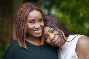 Close-up shot of two Black women embracing outdoors. The woman in the foreground has shoulder-length auburn hair and is wearing a dark-colored top. She has a warm smile, showcasing her slightly crooked teeth, and her expression is one of happiness and affection. The woman behind her has dark brown, straight hair that falls past her shoulders, and she's wearing a light beige, sleeveless top. Her head rests on the shoulder of the woman in front, and she also has a soft smile, conveying a sense of comfort and closeness. The background is softly blurred, showing a green, leafy environment, suggesting a park or garden setting. The focus is sharply on the two women, highlighting their connection and positive emotions.