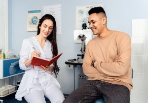 A female doctor talks to a male patient. She holds a notebook. Both are in a medical setting.