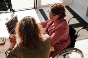 A woman in wheelchair talks to another one. Both are at a table with a laptop on it.