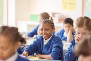 Image of a child in a school uniform sat at a desk with pen in hand. The child smiles at the camera, whilst other children appear blurred in the background, concentrated on reading.